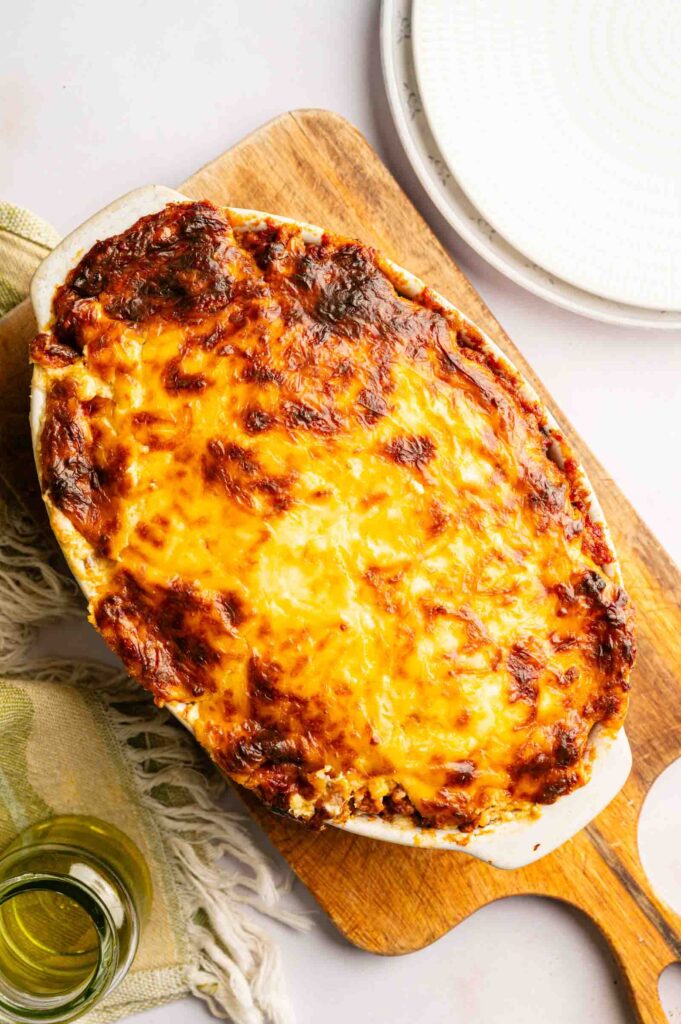 A baked John Wayne Casserole with a golden, browned cheese topping sits on a wooden cutting board next to a plate and a glass of water.