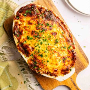 Baked John Wayne Casserole topped with melted, browned cheese and chopped herbs, served in a white dish on a wooden board, with a towel, plates, oil, and serving spoon nearby.