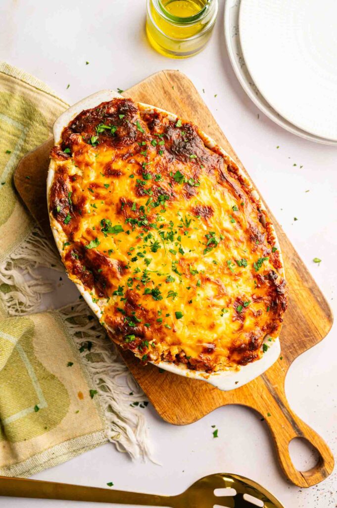 Baked John Wayne Casserole topped with melted, browned cheese and chopped herbs, served in a white dish on a wooden board, with a towel, plates, oil, and serving spoon nearby.