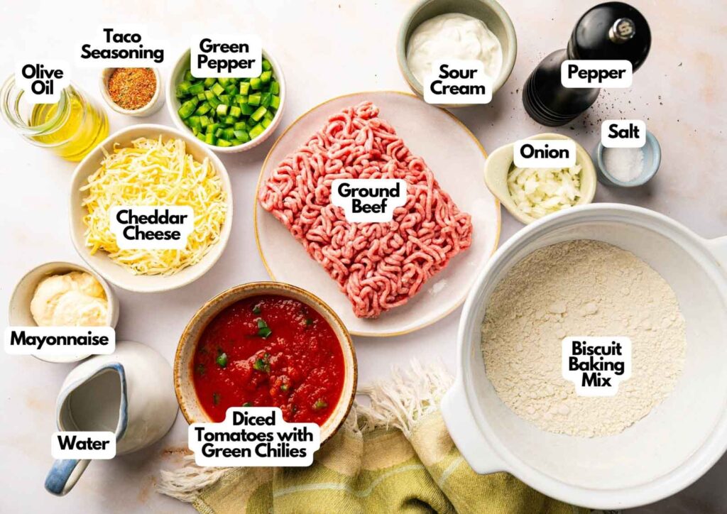 Ingredients for a John Wayne Casserole taco bake are arranged on a table, including ground beef, vegetables, cheese, spices, biscuit mix, and sauces—all clearly labeled with their names.