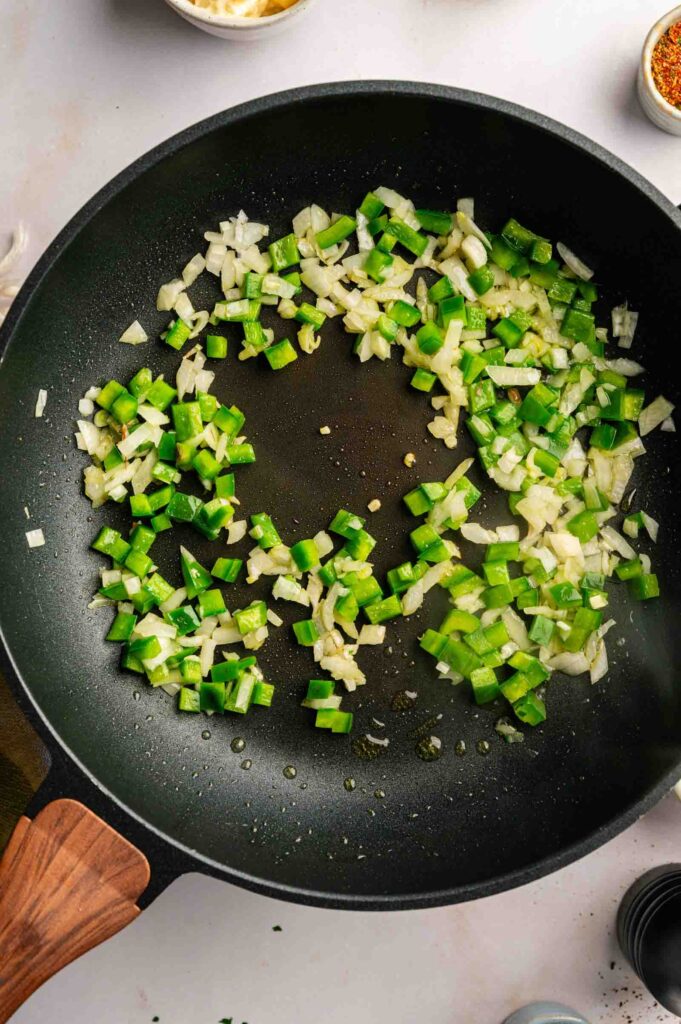 Diced green bell peppers and onions sautéing in a black frying pan with a wooden handle, the perfect start for a hearty John Wayne Casserole.