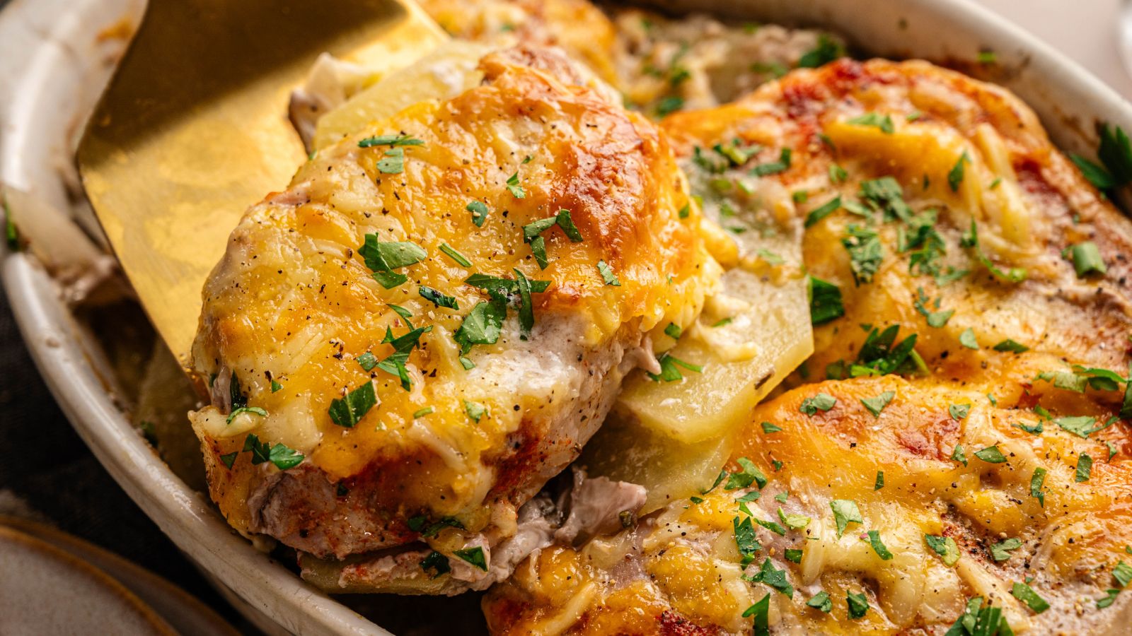 A close-up of a cheesy baked chicken casserole topped with chopped parsley, being served with a spatula.