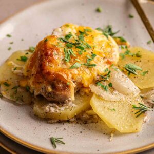 A serving of Pork Chop Casserole topped with melted cheese, placed on thinly sliced potatoes, and garnished with chopped herbs on a ceramic plate.