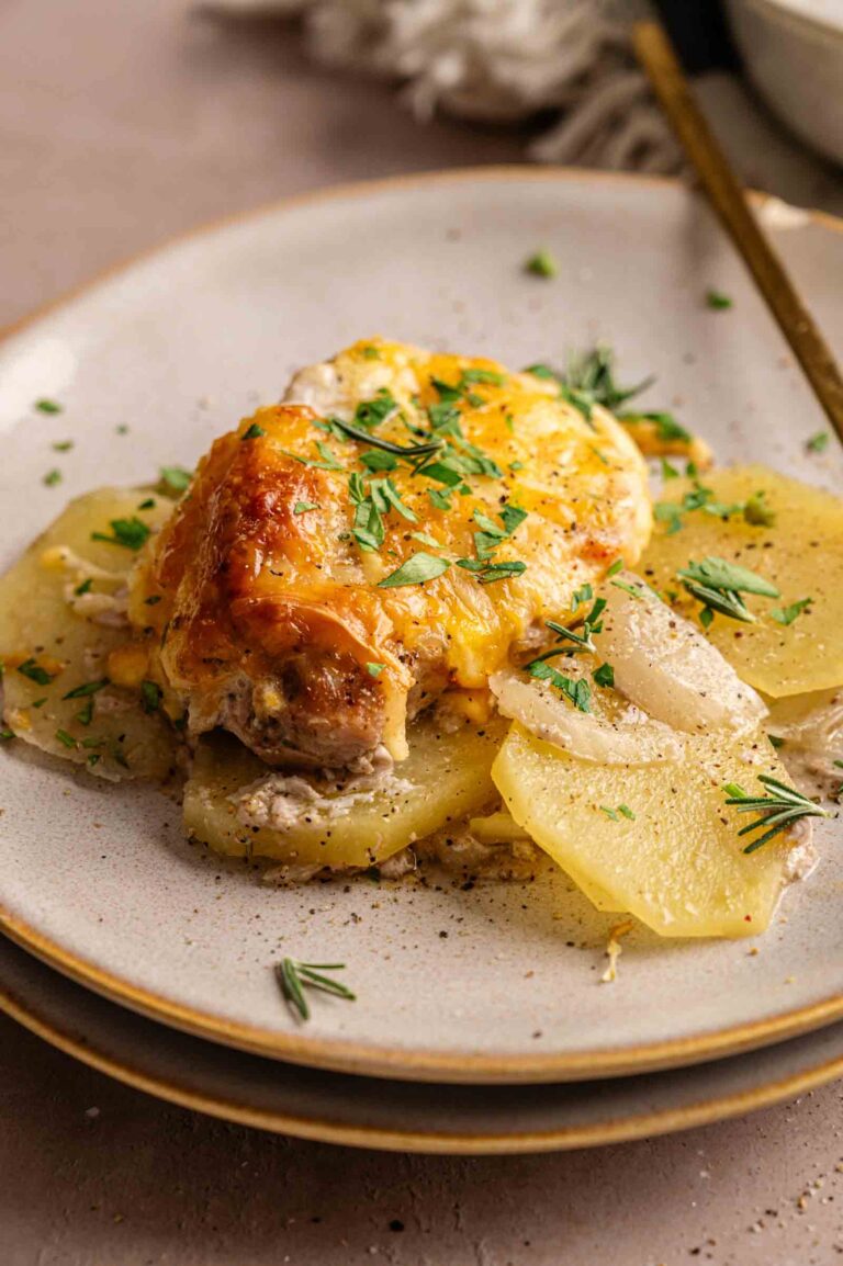 A serving of Pork Chop Casserole topped with melted cheese, placed on thinly sliced potatoes, and garnished with chopped herbs on a ceramic plate.