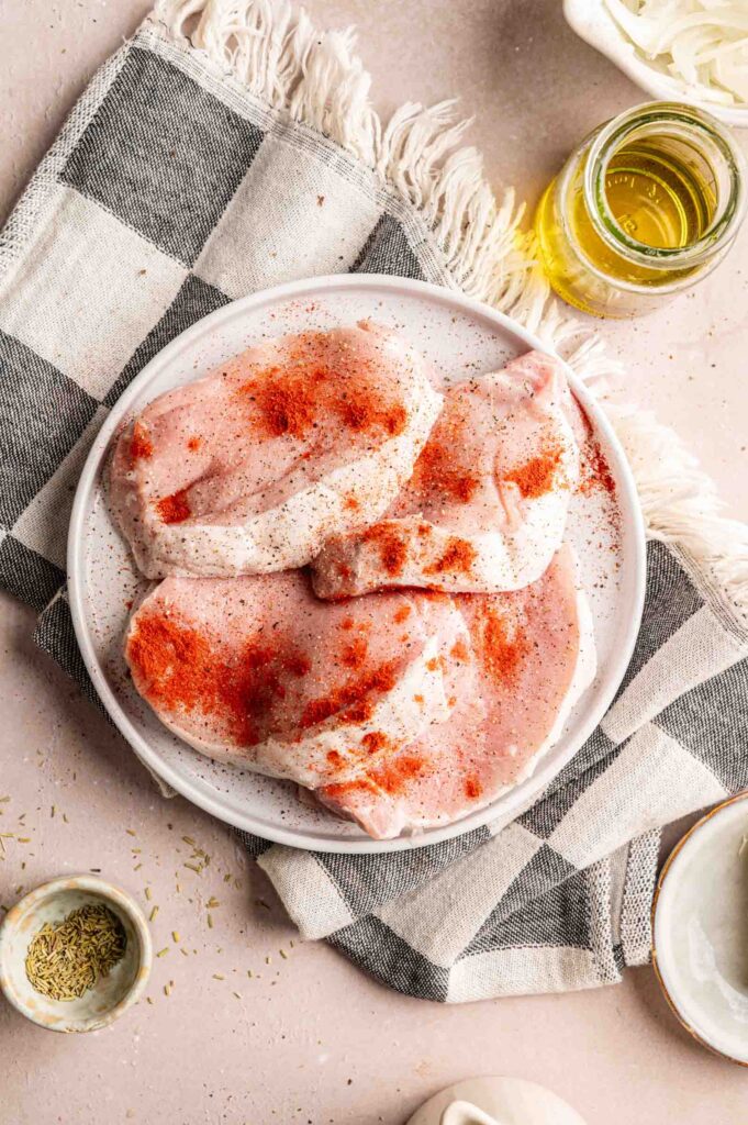 Raw pork chops with seasoning and paprika on a white plate, set on a checkered cloth with a jar of oil and spices nearby&mdash;perfect ingredients to start your Pork Chop Casserole.