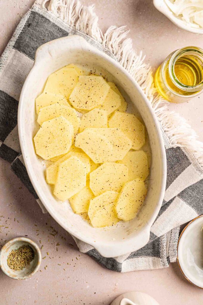 Oval baking dish with a single layer of sliced potatoes seasoned with black pepper, reminiscent of a classic Pork Chop Casserole, placed on a checkered cloth with a jar of oil and spices nearby.