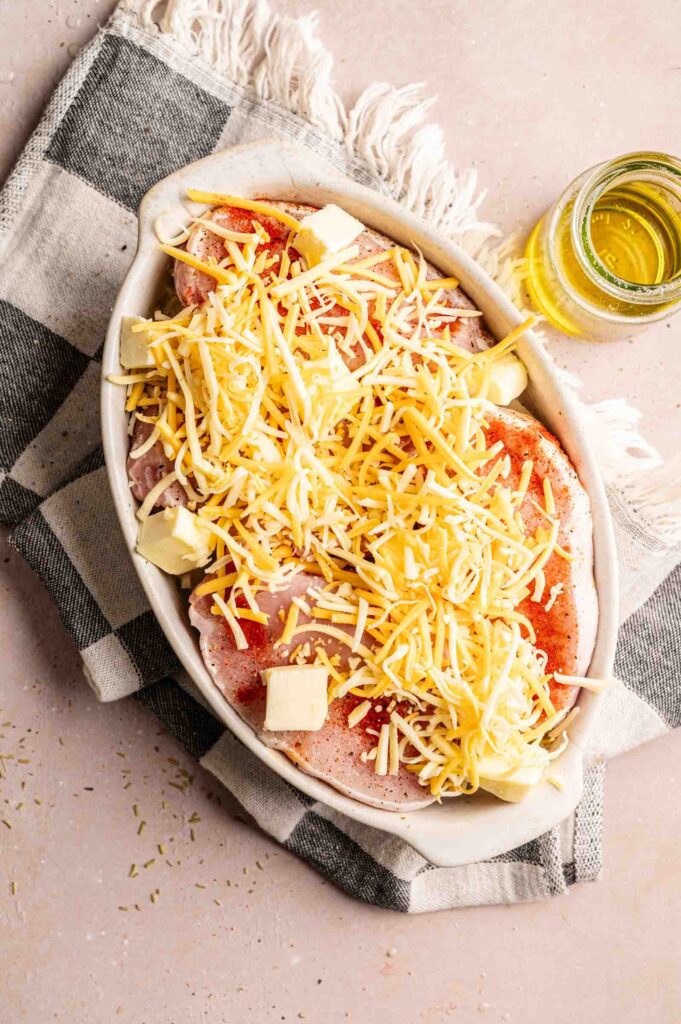 Seasoned raw pork chops layered in a white oval baking dish, topped with a generous mound of shredded cheddar-jack cheese and cubes of butter, placed on a black-and-white checkered napkin next to a small glass jar of olive oil.
