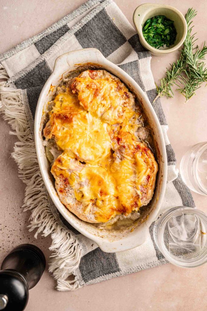 A Pork Chop Casserole with melted cheese on top, placed on a striped cloth, sits next to a bowl of chopped herbs, a sprig of rosemary, two glasses of water, and a pepper grinder.