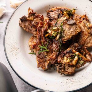 A plate of Air Fryer Lamb Loin Chops garnished with garlic and rosemary, served on a white dish with a black rim.