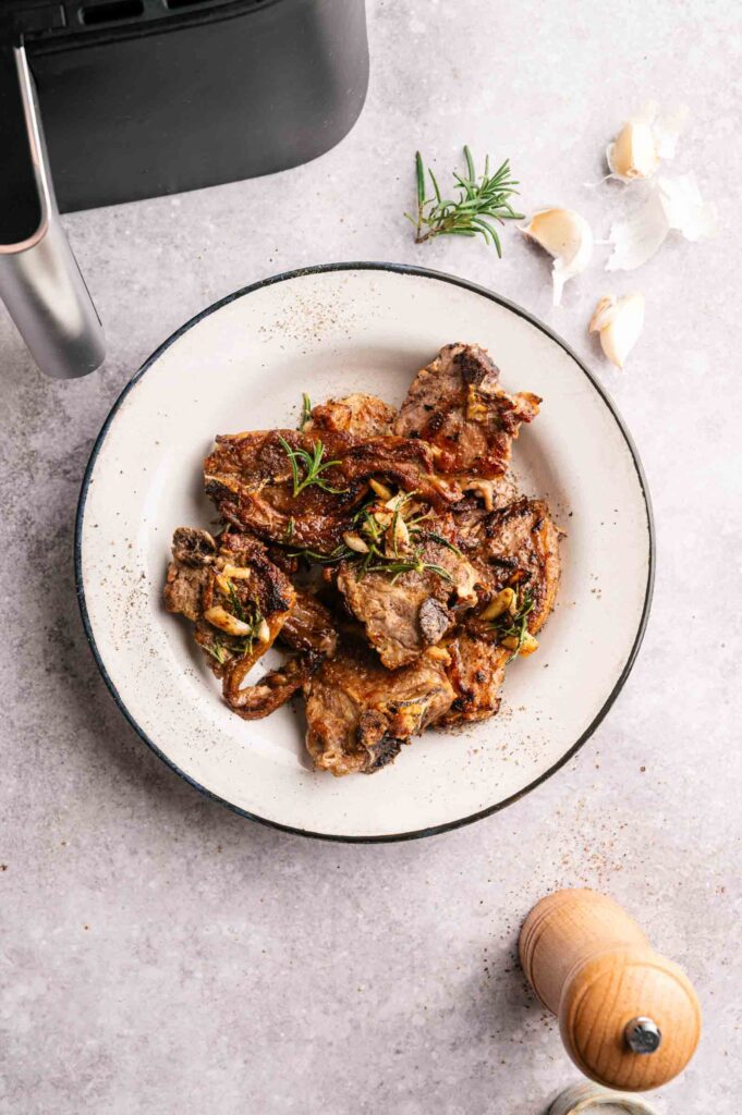 A plate of Air Fryer Lamb Loin Chops garnished with herbs, placed on a light surface next to an air fryer, garlic cloves, rosemary, and a pepper mill.