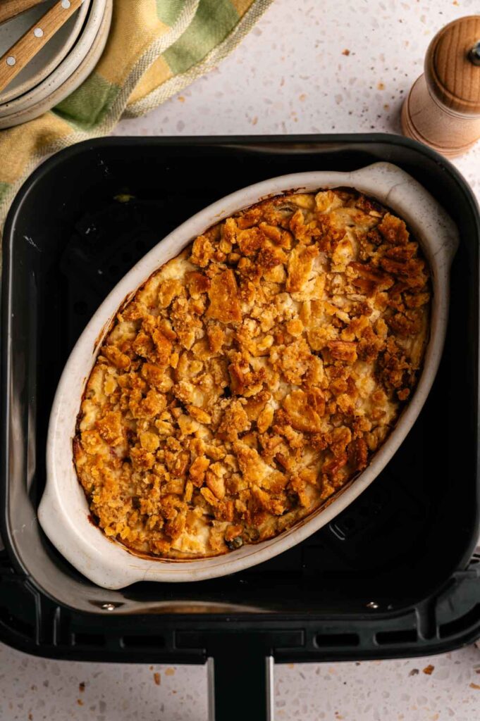 An oval baking dish with a golden, crumb-topped Air Fryer Neiman Marcus Chicken Casserole sits inside an air fryer basket on a speckled countertop.