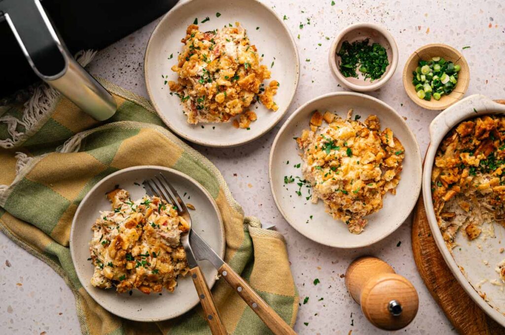 Three plates of Air Fryer Neiman Marcus Chicken Casserole topped with herbs and crispy crumbs, surrounded by chopped green onions, parsley, a pepper grinder, and a dish towel on a light countertop.