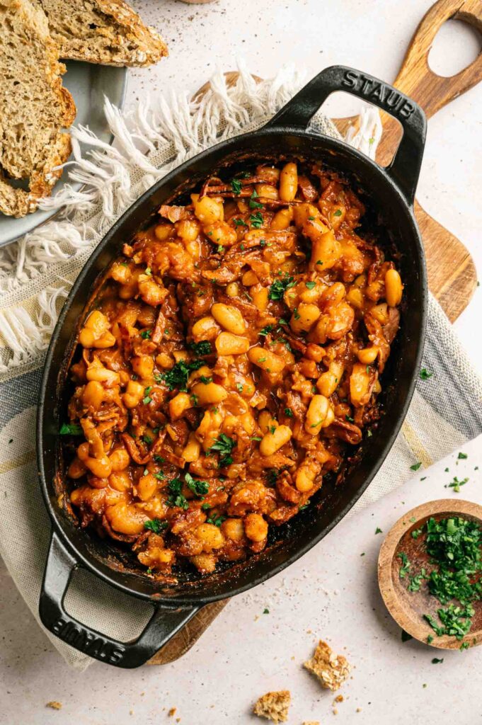 An oval cast iron dish of Baked Beans in a rich tomato-based sauce, garnished with chopped parsley, sits on a cloth with sliced bread nearby.