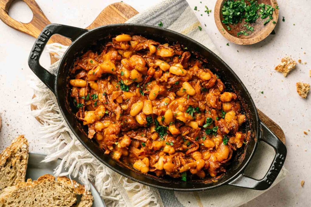 An oval casserole dish filled with Baked Beans in tomato sauce, garnished with chopped herbs, sits on a table next to bread slices and a small bowl of herbs.