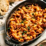 A dish filled with classic baked beans in a rich tomato-based sauce, garnished with chopped herbs. Slices of bread are visible in the background.