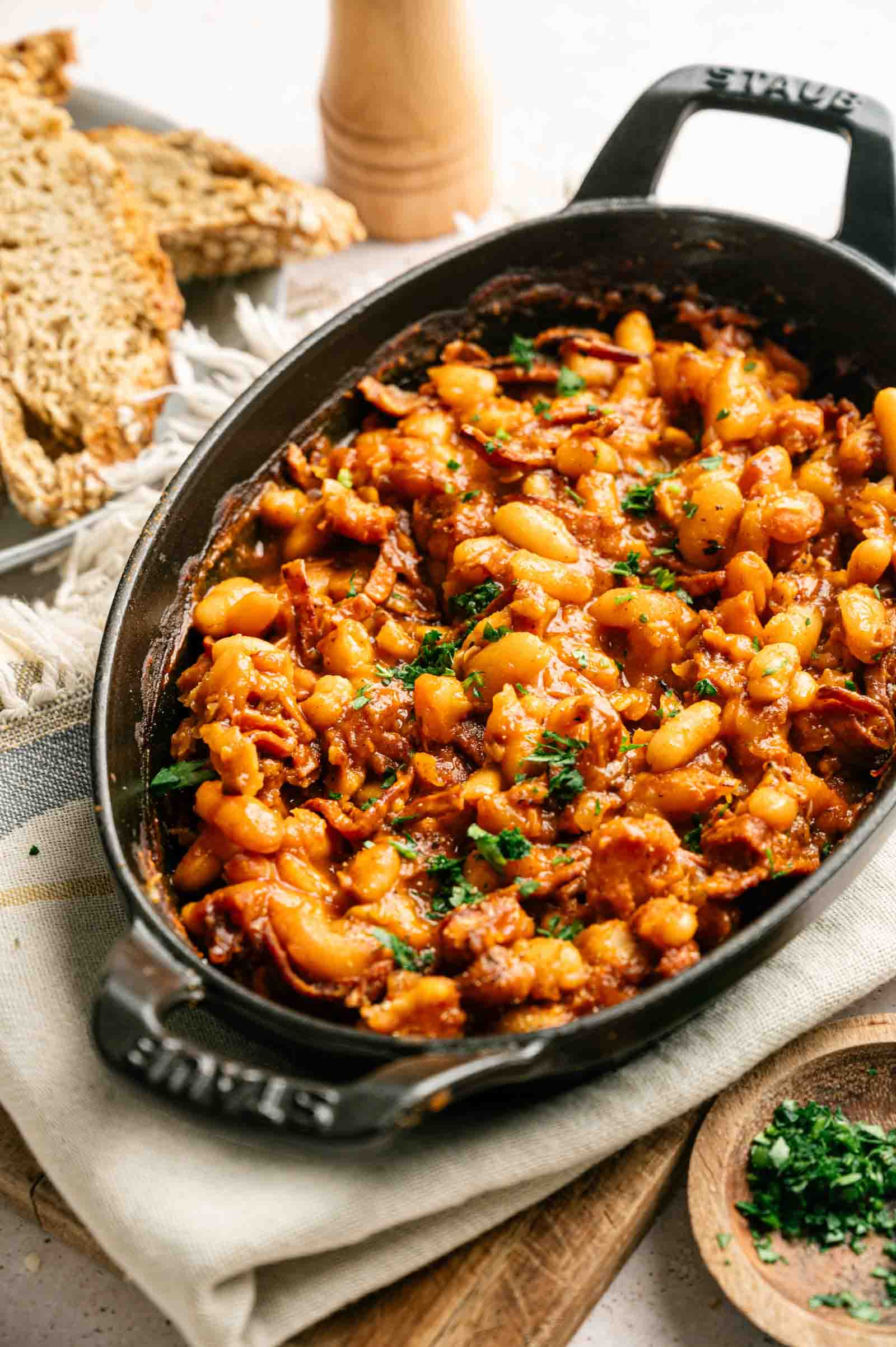 A dish filled with classic baked beans in a rich tomato-based sauce, garnished with chopped herbs. Slices of bread are visible in the background.