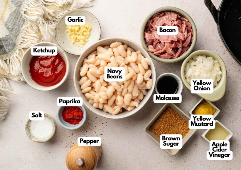 Bowls containing ingredients for Baked Beans—navy beans, ketchup, paprika, salt, pepper, garlic, bacon, molasses, yellow onion, mustard, brown sugar, and apple cider vinegar—are arranged on a table.