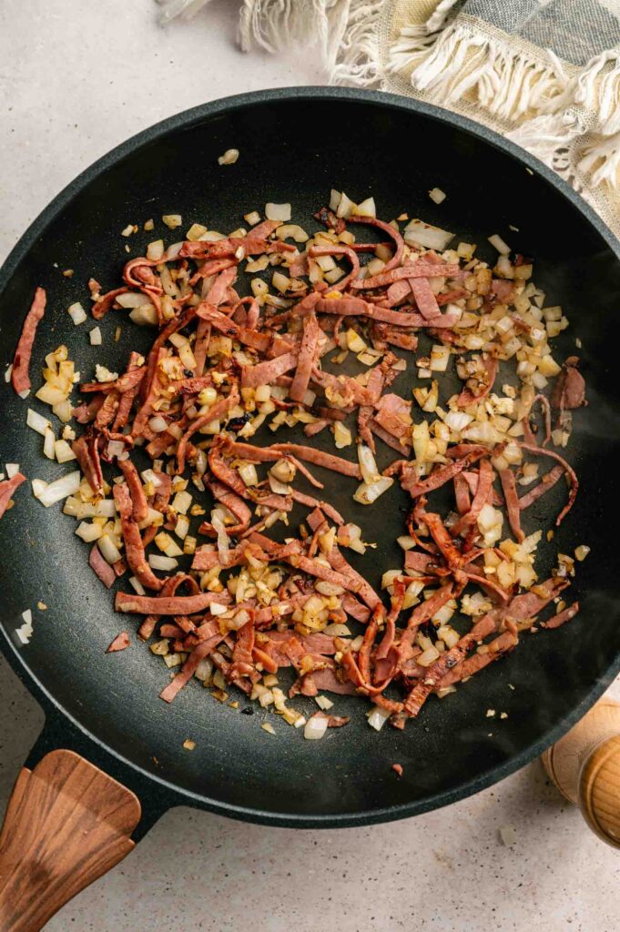 Sliced cooked meat and diced onions are sautéing in a black frying pan on a light-colored countertop, ready to be served with hearty baked beans.