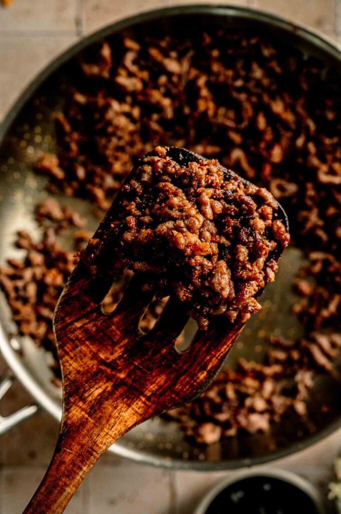 A wooden spatula holds cooked, browned ground beef over a skillet filled with more meat, ready to be added to a zesty Doritos Taco Salad.