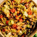 A bowl of Doritos Taco Salad with lettuce, cherry tomatoes, black beans, cheese, and crunchy tortilla chips, mixed with wooden utensils on a tiled surface.
