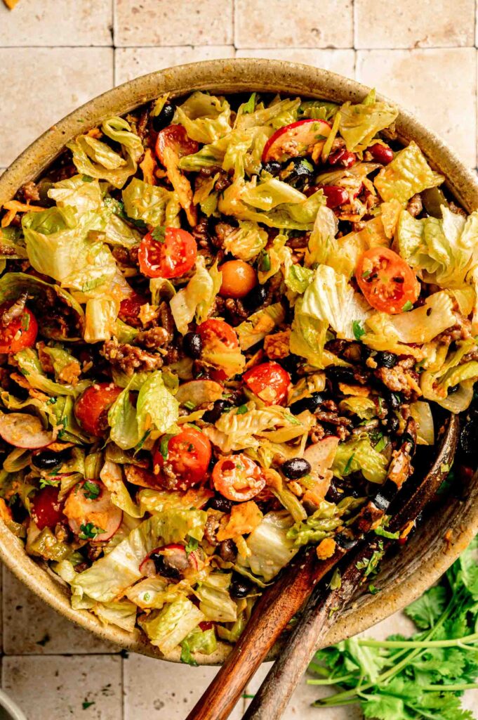 A bowl of Doritos Taco Salad with lettuce, cherry tomatoes, black beans, cheese, and crunchy tortilla chips, mixed with wooden utensils on a tiled surface.