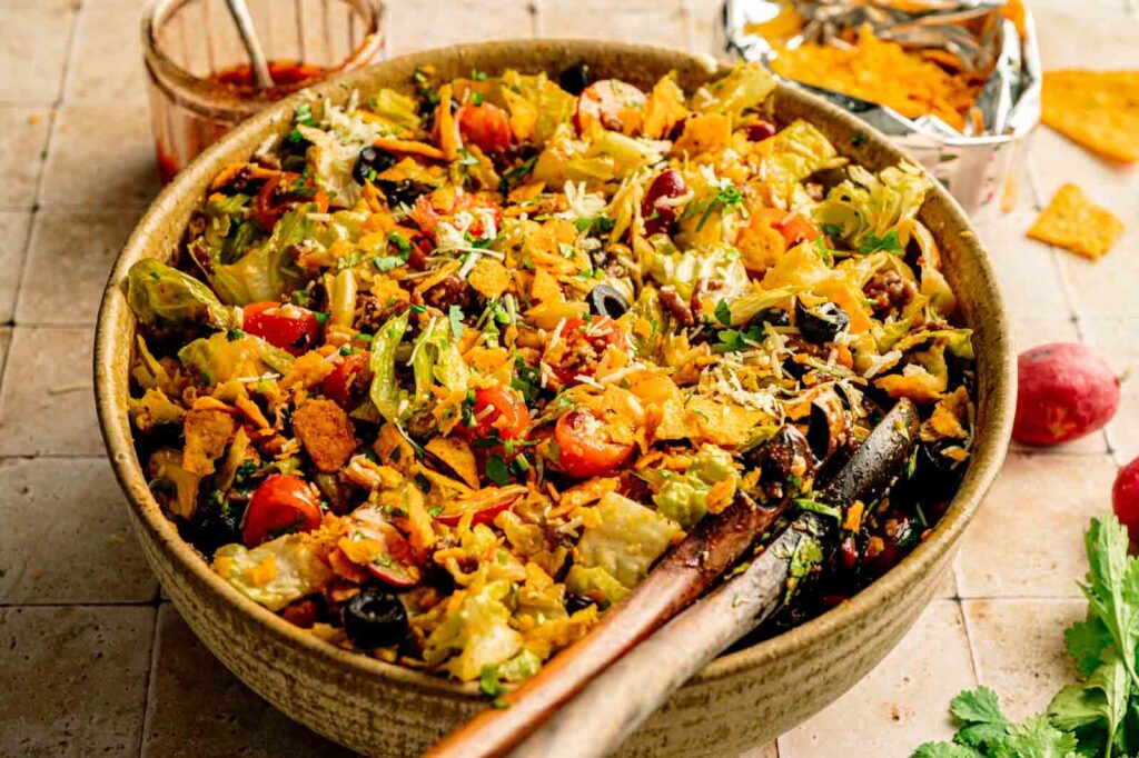 A bowl of Doritos Taco Salad with lettuce, cherry tomatoes, shredded cheese, olives, and crushed tortilla chips, with salad tongs resting inside.