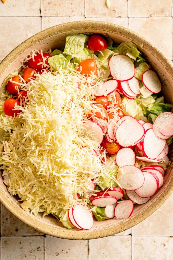 A large bowl filled with chopped romaine lettuce, cherry tomatoes, sliced radishes, and shredded cheddar cheese as the base for Doritos Taco Salad.