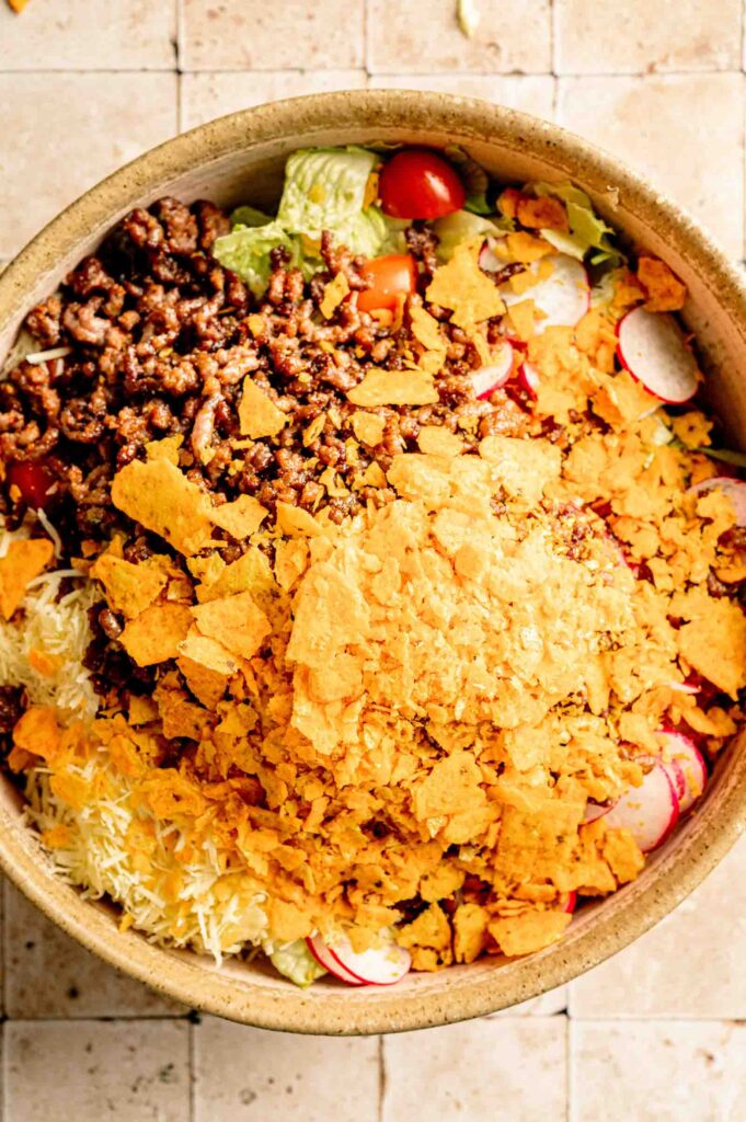 A large bowl of Doritos Taco Salad base topped with seasoned ground beef and crushed Nacho Cheese Doritos, ready to be tossed together.