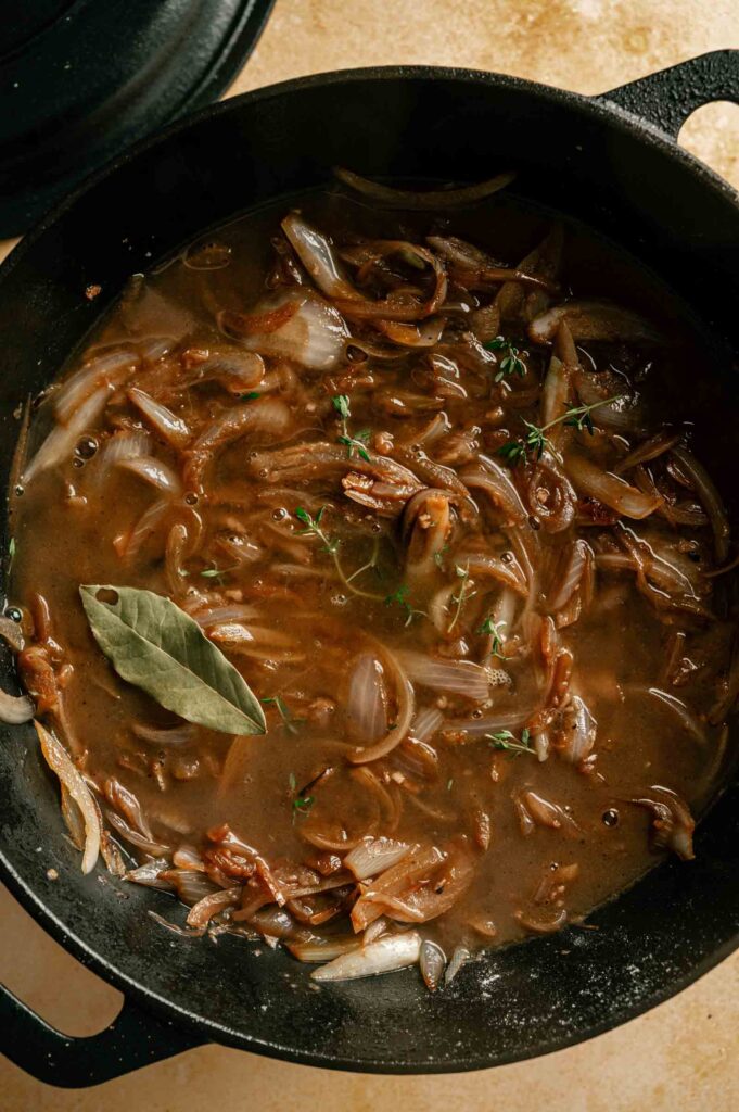 A black pot filled with sliced onions, brown broth, fresh thyme, and a bay leaf—perfect for inspiring your next French Onion Pot Roast.