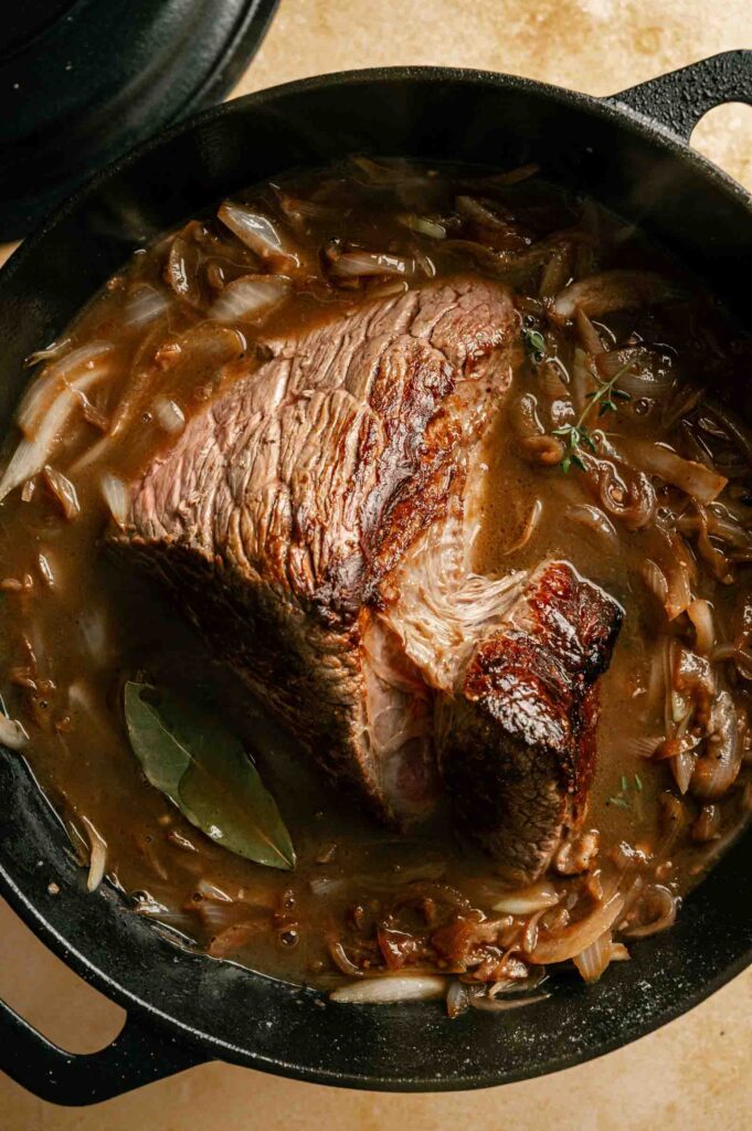 A large piece of French Onion Pot Roast sits in a pot with brown gravy, sliced onions, a bay leaf, and fresh herbs.