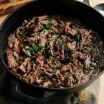 French Onion Pot Roast shredded beef with sauce and chopped herbs in a black cast iron pot, with a cutting board and knife visible in the background.