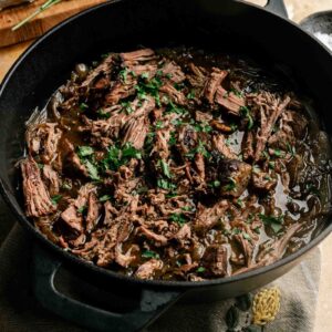 French Onion Pot Roast shredded beef with sauce and chopped herbs in a black cast iron pot, with a cutting board and knife visible in the background.