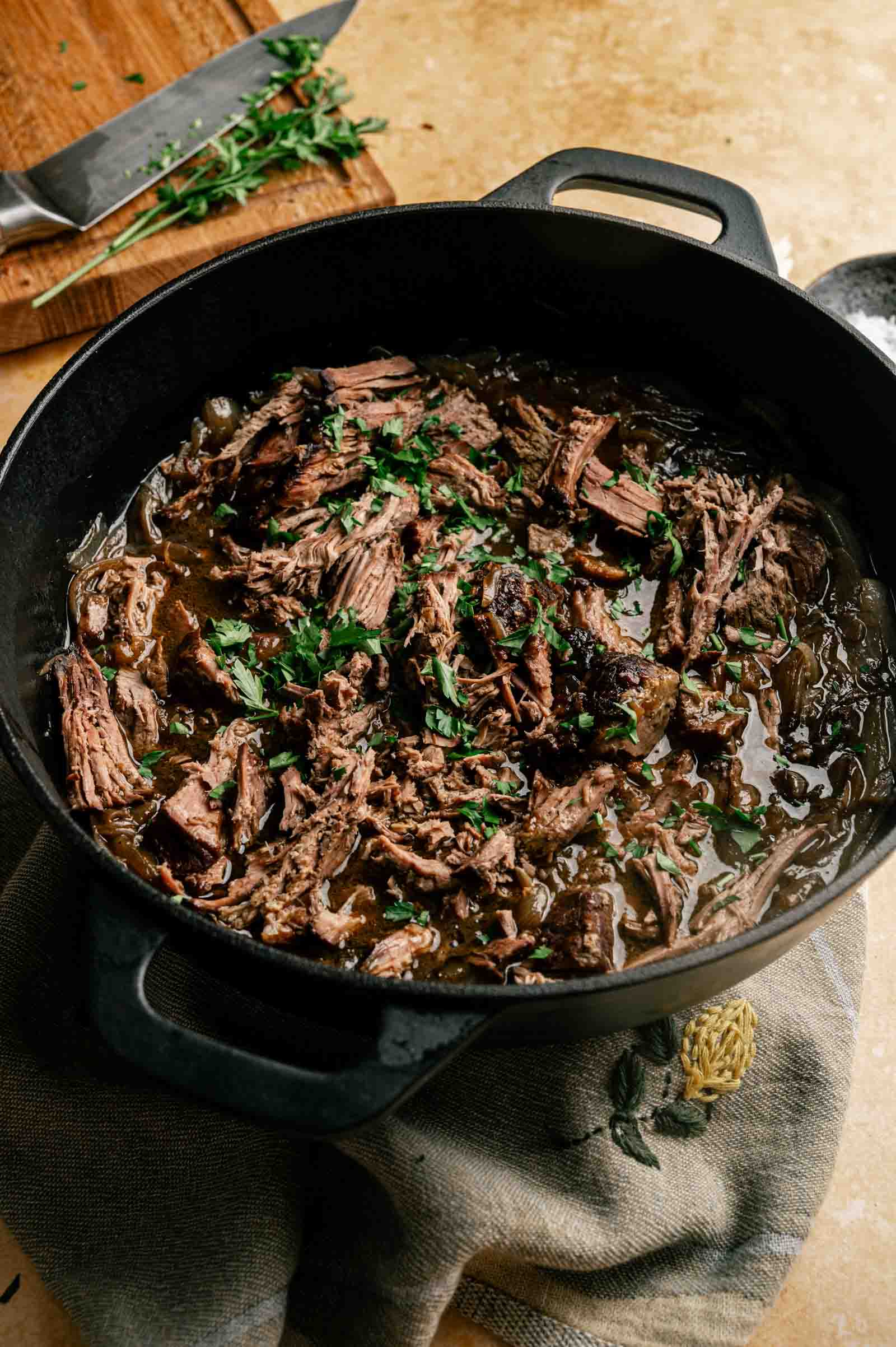 French Onion Pot Roast shredded beef with sauce and chopped herbs in a black cast iron pot, with a cutting board and knife visible in the background.