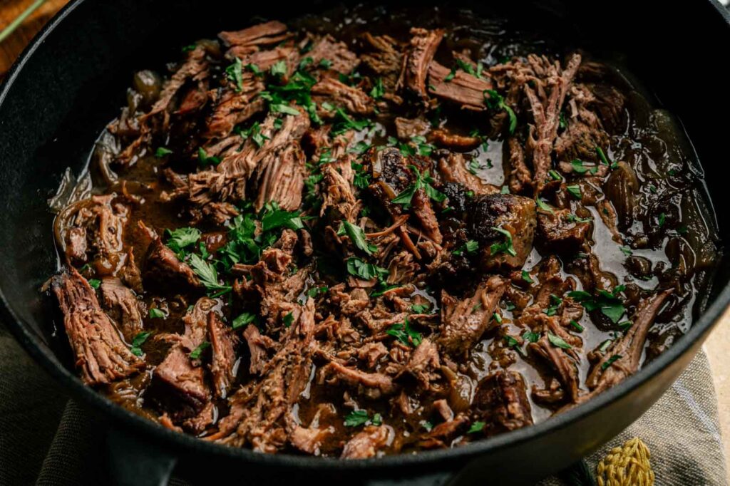 French Onion Pot Roast: Shredded pot roast with herbs in a rich, dark sauce, served in a black cast iron pot and garnished with chopped parsley.