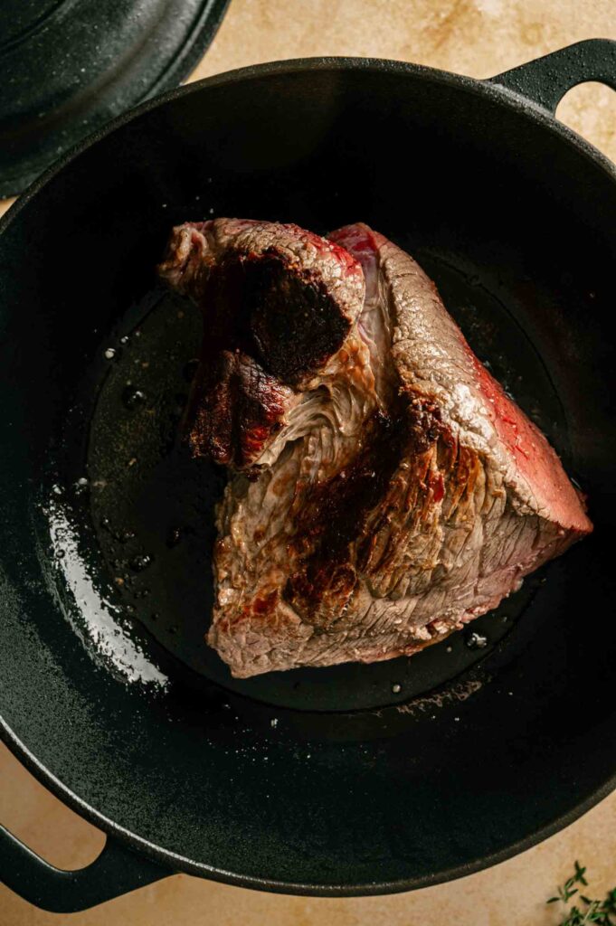 A seared piece of chuck roast sits in a black cast iron skillet, showing browned edges and some visible fat.