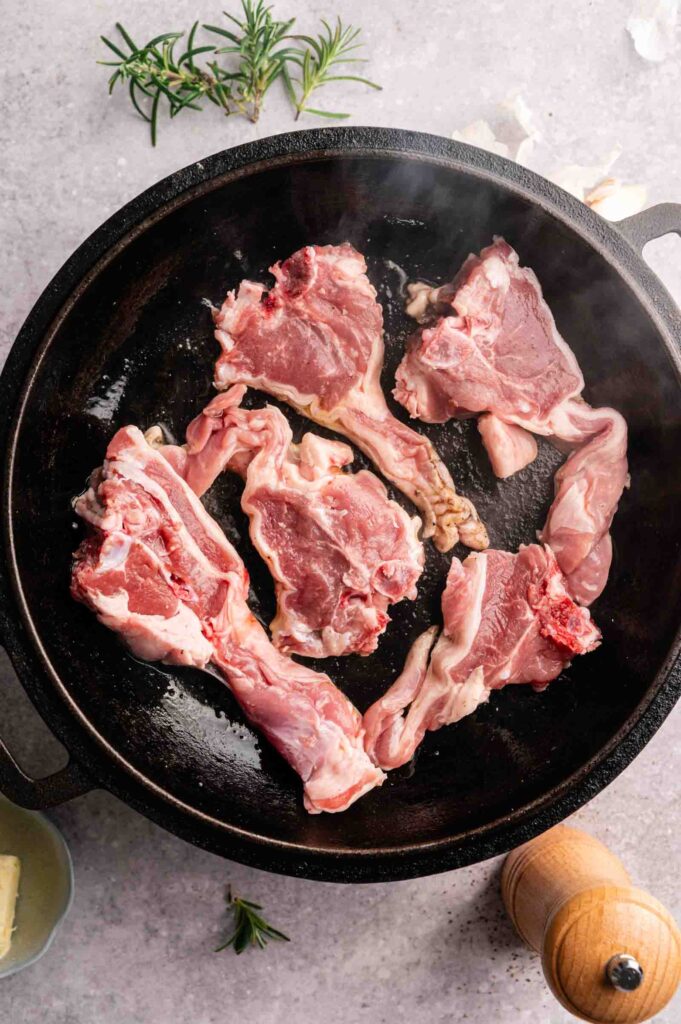 Raw lamb chops arranged in a black cast iron skillet, with a sprig of rosemary and a wooden pepper grinder nearby—perfect for preparing Air Fryer Lamb Loin Chops on a light countertop.