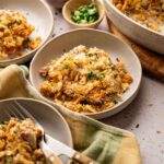 Three bowls of Air Fryer Neiman Marcus Chicken Casserole topped with crispy crumbs, served with forks, alongside a casserole dish and small bowls of chopped green onions and parsley.