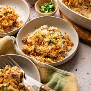 Three bowls of Air Fryer Neiman Marcus Chicken Casserole topped with crispy crumbs, served with forks, alongside a casserole dish and small bowls of chopped green onions and parsley.