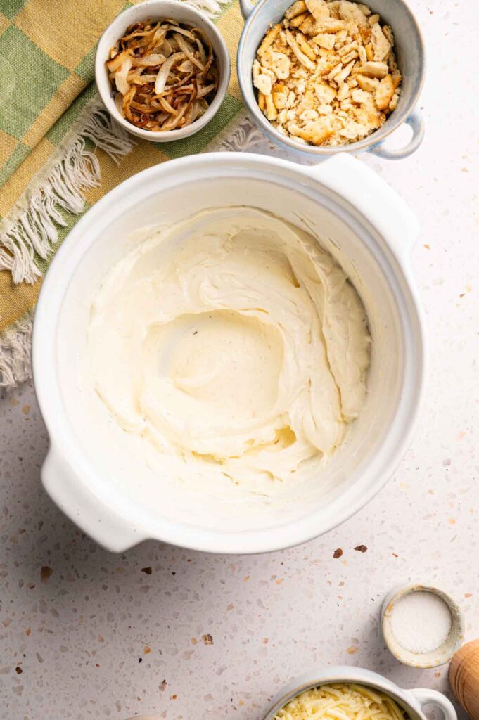A mixing bowl filled with creamy blended base sits on a countertop, alongside toppings for Air Fryer Neiman Marcus Chicken Casserole like crispy onions, crushed crackers, and shredded cheese in small bowls.