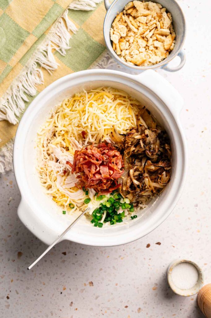 A white bowl holds ingredients for Air Fryer Neiman Marcus Chicken Casserole: shredded cheese, cooked bacon bits, saut&eacute;ed mushrooms, chopped green onions, and shredded chicken. Crushed crackers are in a separate bowl on the countertop.
