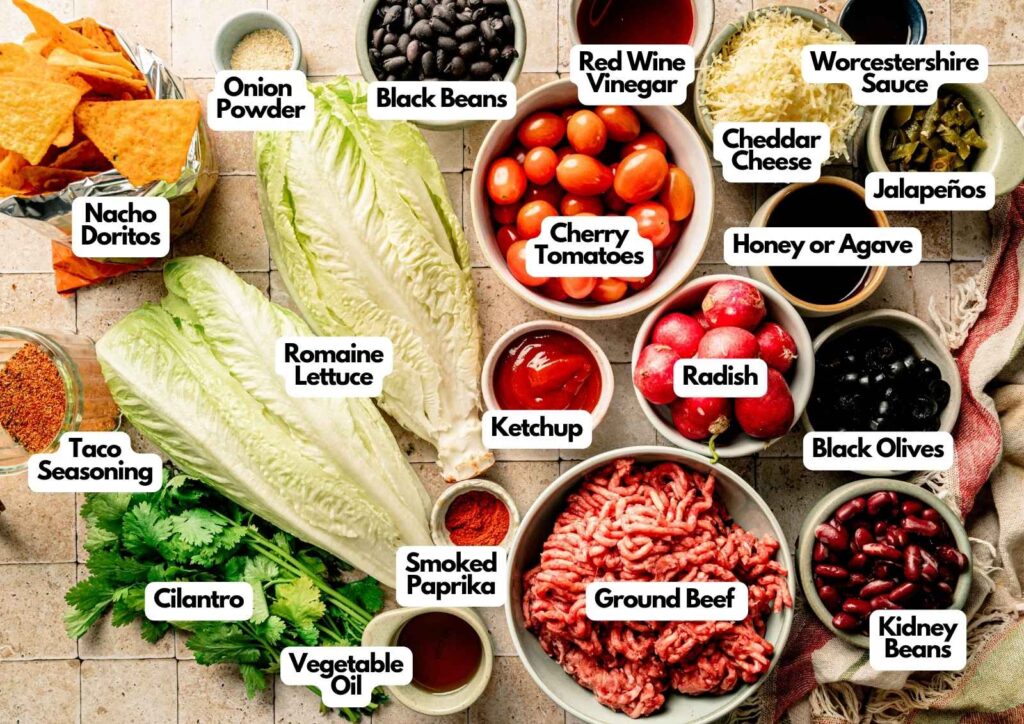 A variety of taco ingredients, including ground beef, lettuce, beans, vegetables, seasonings, sauces, and chips are arranged on a countertop for a delicious Doritos Taco Salad, each labeled with its name.